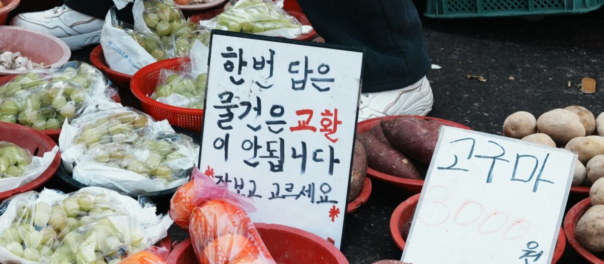 전통시장 좌판의 빨간 바구니에 담긴 샤인머스캣과 고구마, 감자, 그리고 손글씨 가격표
