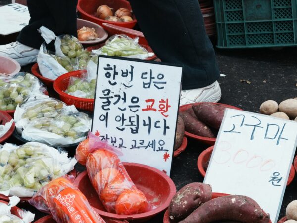 전통시장 좌판의 빨간 바구니에 담긴 샤인머스캣과 고구마, 감자, 그리고 손글씨 가격표